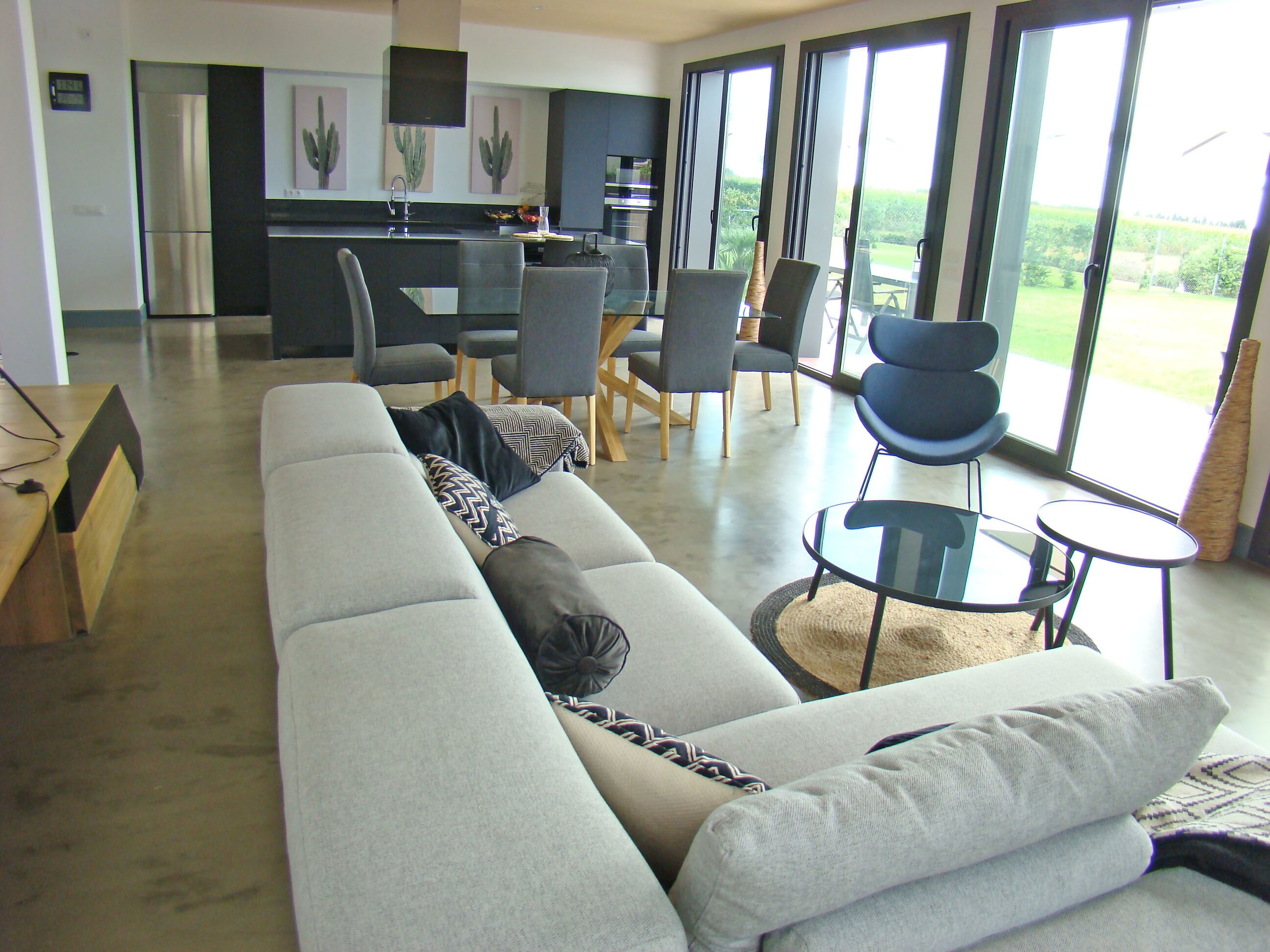 A modern living room with a sofa, chairs, a glass coffee table, and a kitchen area with a refrigerator, sink, and dining table.