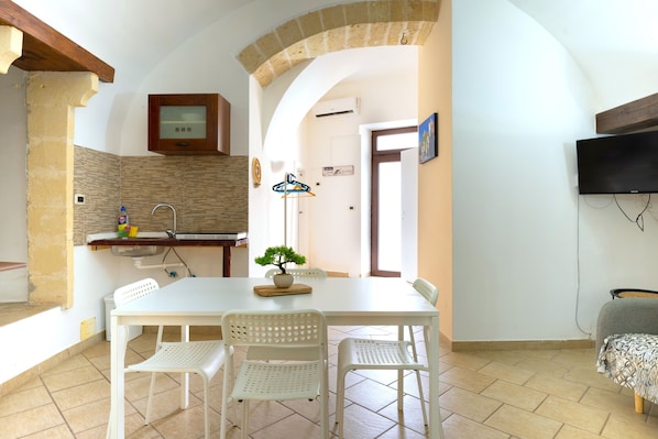 A dining area with a white table and chairs, a small kitchen with a sink and cabinets, a potted plant, and a television mounted on the wall. - Apartment 'Sg  Family' with Air Conditioning (Massafra)