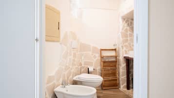 A bathroom with a stone wall, a bidet, a toilet, and a wooden shelf.