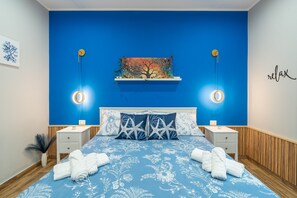 Ein Schlafzimmer mit blauer Wand, einem Bett mit weißer Blumenbettwäsche, zwei Nachttischen mit Lampen, einem Baumgemälde und einem Schild mit der Aufschrift 'relax'.