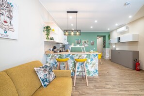 Eine moderne Küche mit einer zentralen Kochinsel, ein Essbereich mit gelben Stühlen und ein Wohnzimmer mit einem senffarbenen Sofa.
