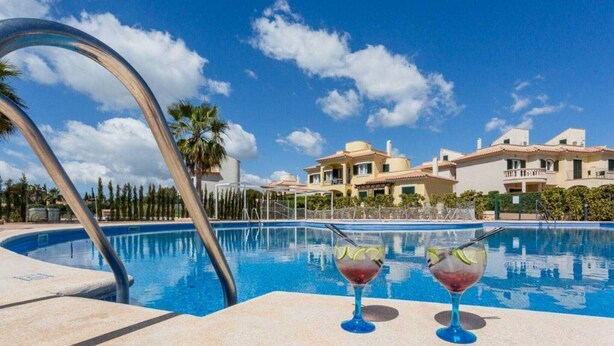 A swimming pool with two drinks on the edge, surrounded by a clear blue sky and a large building in the background. - Holiday Home 'Residence Club El Paraiso 30' with Sea View, Wi-Fi and Air Conditioning (Sa Ràpita)