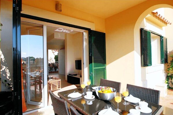 Un área de comedor al aire libre con una mesa preparada para dos personas, un tazón de fruta y un vaso de jugo de naranja.