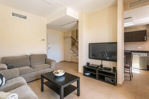Un salon comprenant un canapé, une table basse avec un saladier, un téléviseur et un coin cuisine avec un lave-vaisselle.