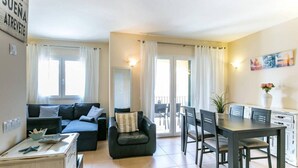 Un salón con sofá, sillón, mesa de comedor y sillas. Tiene un balcón con barandilla y vistas al exterior.