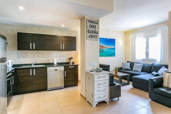 A modern kitchen with dark cabinets, a dishwasher, and a coffee maker. There is a dining area with a sofa and a small table. A sign with text is on the wall, and a painting is above the sofa.