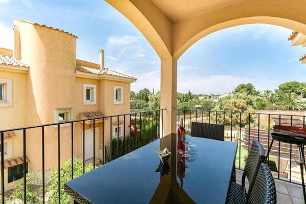 A balcony with a table and chairs, a bottle of wine, and a view of a building and greenery.