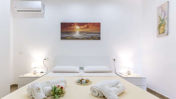 Une chambre d’hôtel avec un lit, deux tables de chevet, un climatiseur, un tableau représentant un coucher de soleil et un vase avec des fleurs.