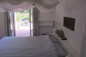 Un dormitorio con cama, mesita de noche, lámpara, estante con libros y vistas a través de un ventanal.
