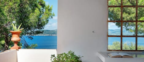 Un balcon donnant sur la mer, une plante en pot et une fenêtre avec vue sur les arbres.