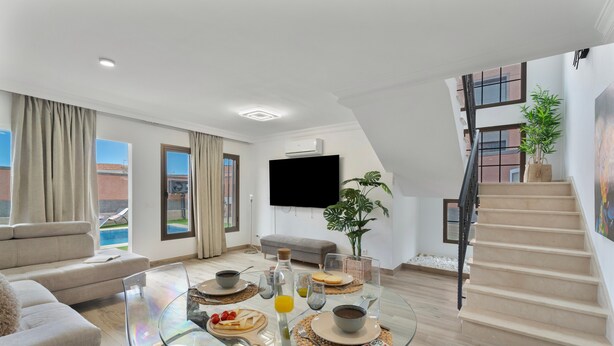 A modern living room with a glass dining table, a sofa, a staircase, and a view of a building outside.
