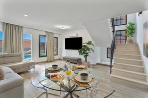 A modern living room with a glass dining table, a sofa, a staircase, and a view of a building outside.