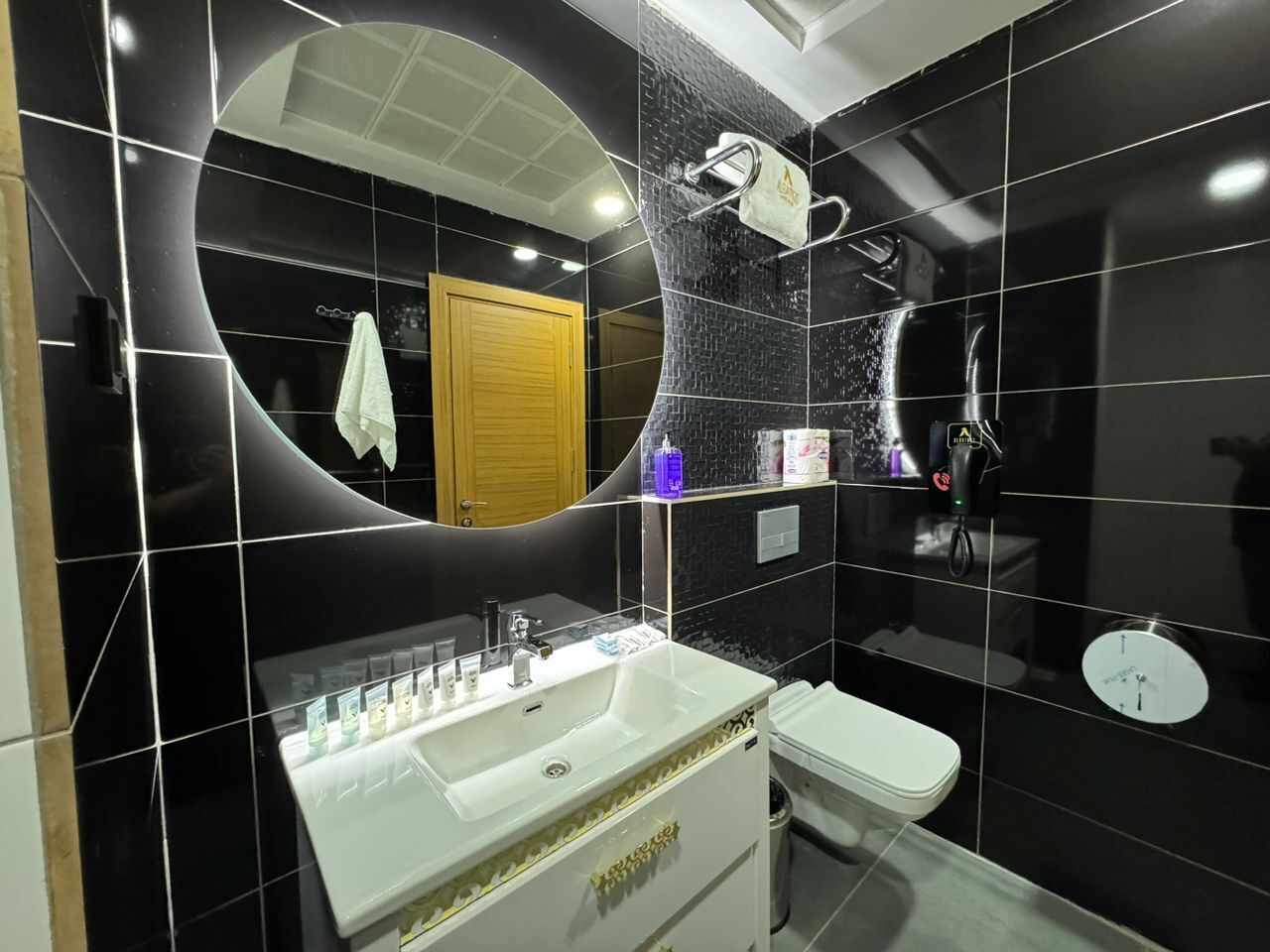 Un baño moderno con un espejo curvo, un lavamanos blanco con detalles dorados, un inodoro y una zona de ducha.
