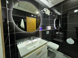 Un baño moderno con un espejo curvo, un lavamanos blanco con detalles dorados, un inodoro y una zona de ducha.