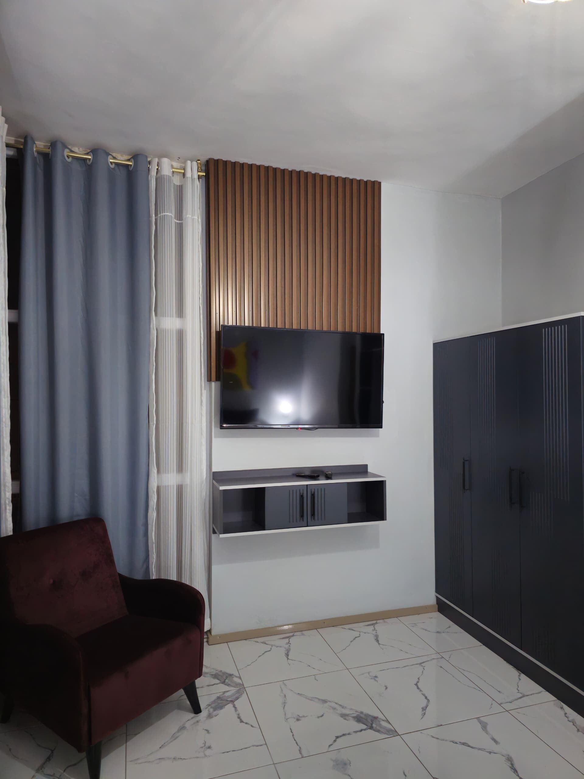 Una sala de estar con un televisor de pantalla plana montado en la pared, un armario de color oscuro, un sofá rojo y una cortina con un velo translúcido.