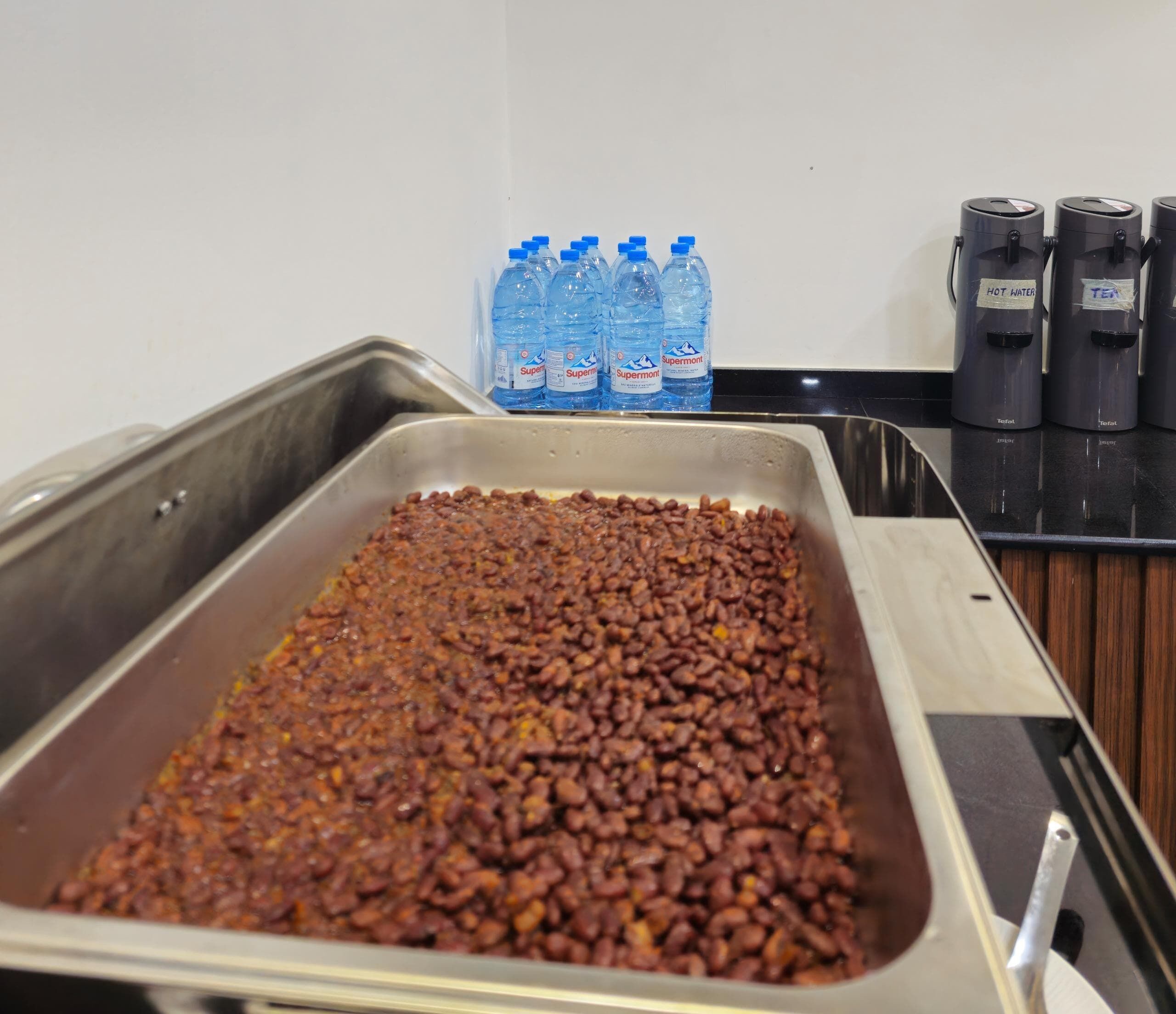 A buffet area with a large tray of cooked beans, several water bottles, and coffee machines.