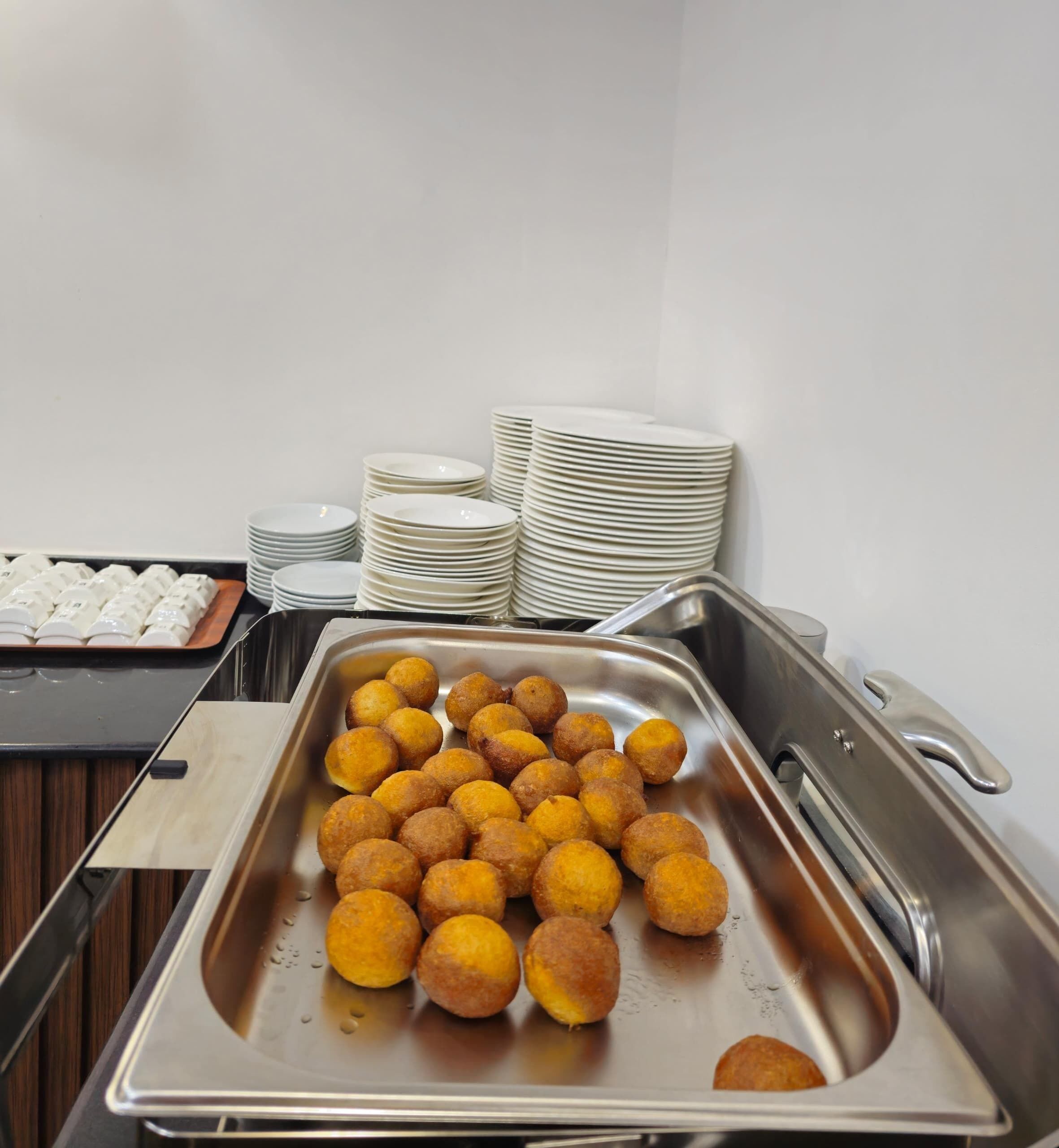 A buffet setup with a tray of golden-brown fried items, stacked white plates, and a metal serving tray.