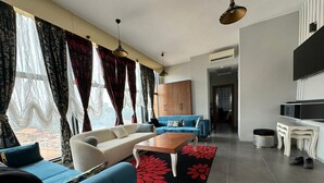 A modern living room with a sofa, a coffee table, and a TV. There is a window with curtains and a view outside. - Albatros Premium Hôtel (Yaoundé)