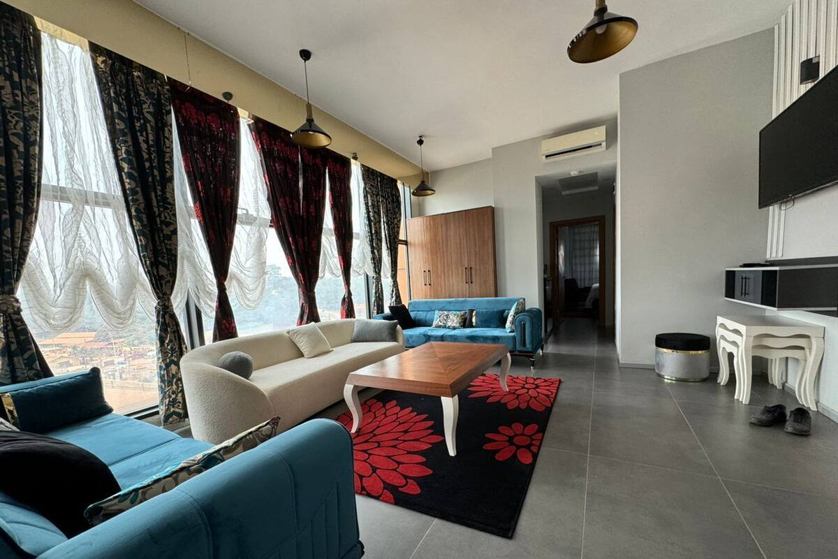 A modern living room with a sofa, a coffee table, and a TV. There is a window with curtains and a view outside.