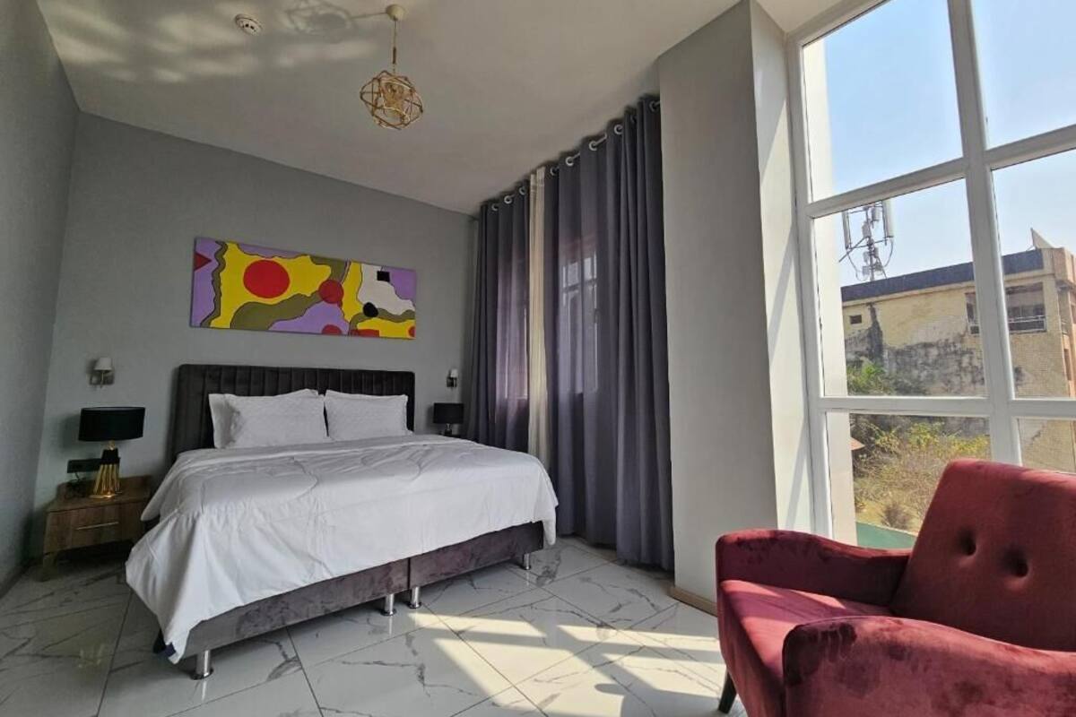 A bedroom with a bed, a red chair, a painting, and a large window.