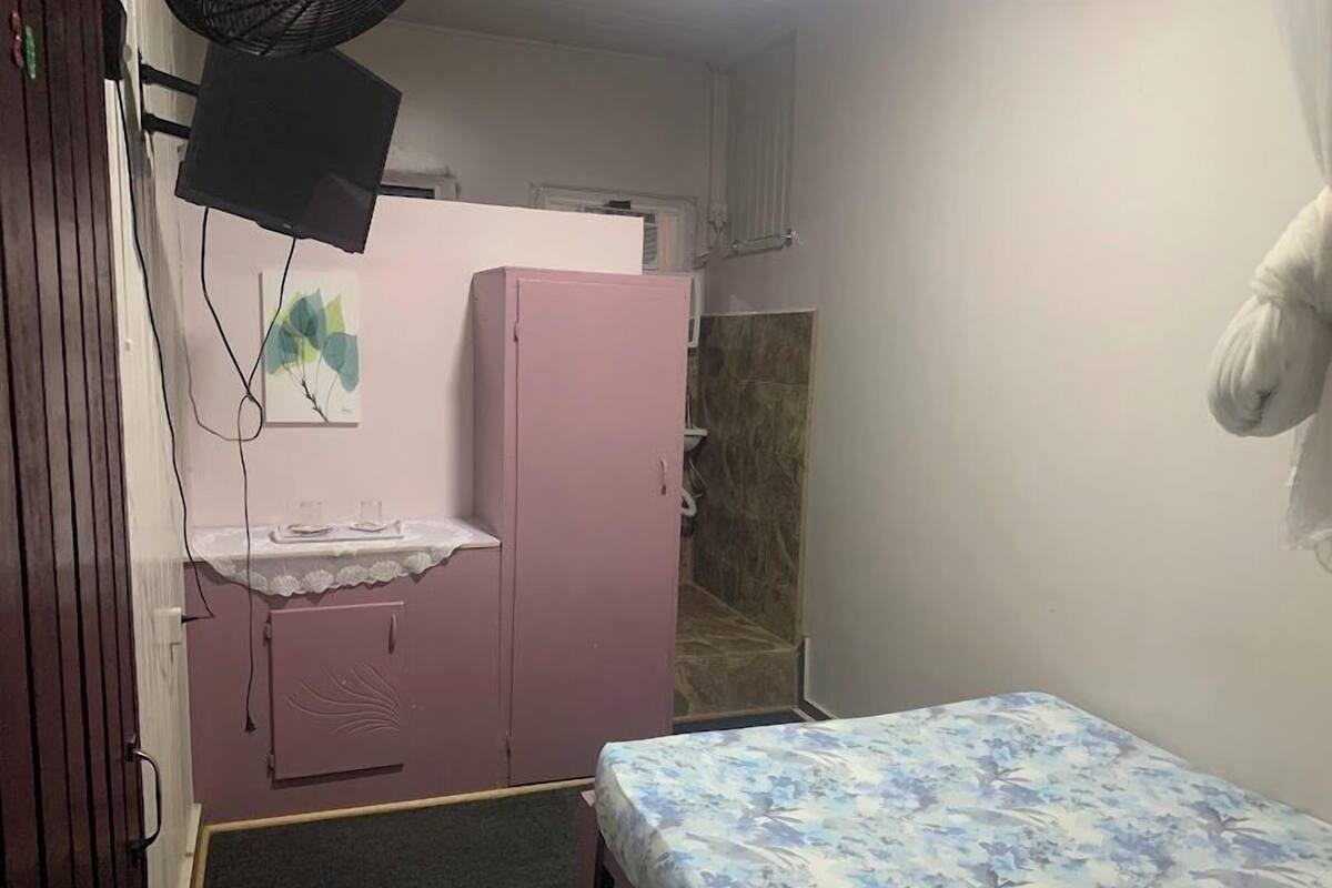 A small room with a pink wardrobe, a bed with a floral bedspread, a small table with a white lace tablecloth, and a wall-mounted fan.