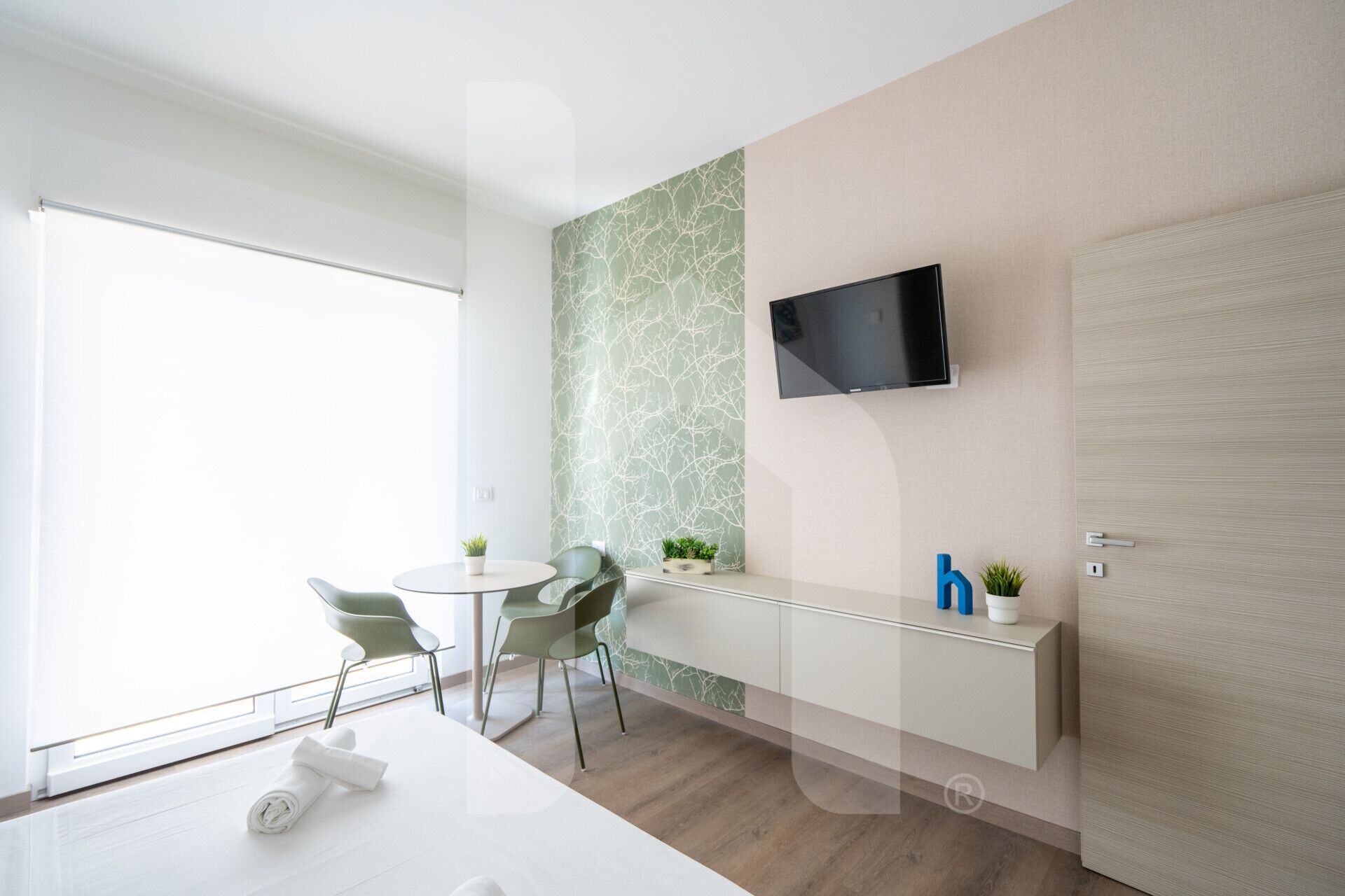 A modern hotel room with a large window, a flat-screen TV mounted on the wall, a small round table with chairs, and a wooden floor.