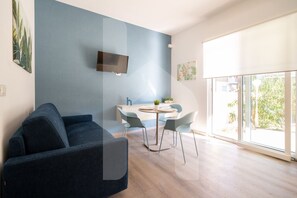 A modern living room with a sofa, a dining table with chairs, a flat-screen TV mounted on the wall, and large windows providing natural light.