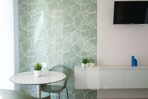 A modern room with a round table, green chairs, a white cabinet with plants, and a flat-screen TV mounted on the wall.