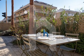 An outdoor dining area with a table set for two, surrounded by chairs, with a building in the background.