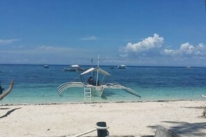 A beach with a boat docked on the shore, clear blue water, and several boats in the distance. - Blue Ocean Bohol Beachfront (Panglao Municipality)