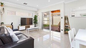 Una sala moderna con un televisor de pantalla plana, un sofá de cuero negro, una mesa de centro blanca y una zona de comedor con sillas blancas.