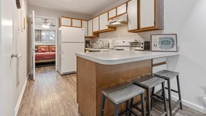 A compact kitchen with wooden cabinets, a central island with stools, a built-in oven, and a refrigerator. - The Ward - Resort Style Gameday Condo (Tallahassee)