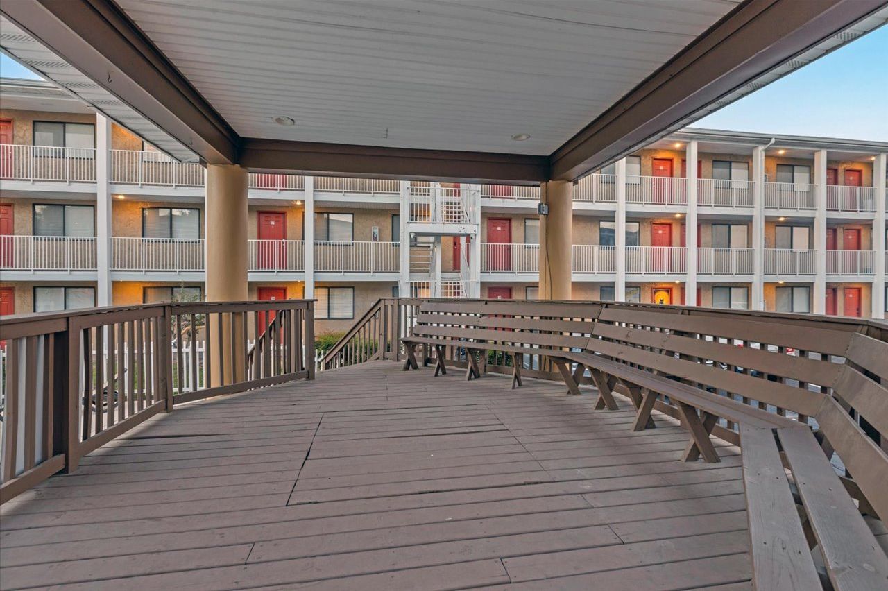 A covered outdoor area with wooden benches and railings, leading to a multi-story building with balconies and red doors.