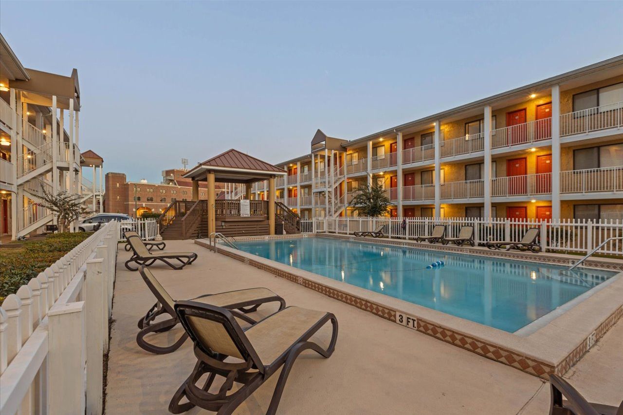 A hotel with a swimming pool, lounge chairs, and a white fence.