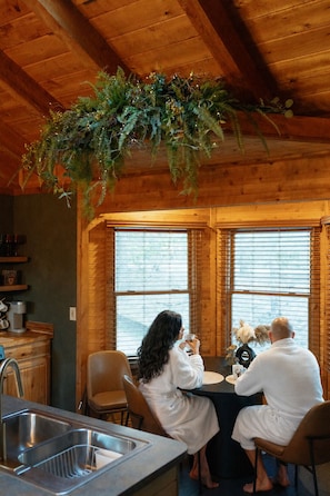 Ein rustikales Holz-Blockhaus-Interieur mit Esstisch, zwei Personen in Bademänteln, einer hängenden Pflanze und einer Küchenzeile.