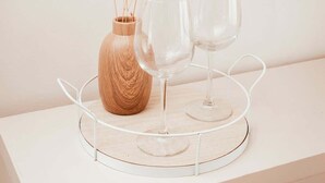 Une table blanche sur laquelle repose un plateau rond en fil de fer, contenant un verre transparent et un vase en bois orné de fleurs séchées.