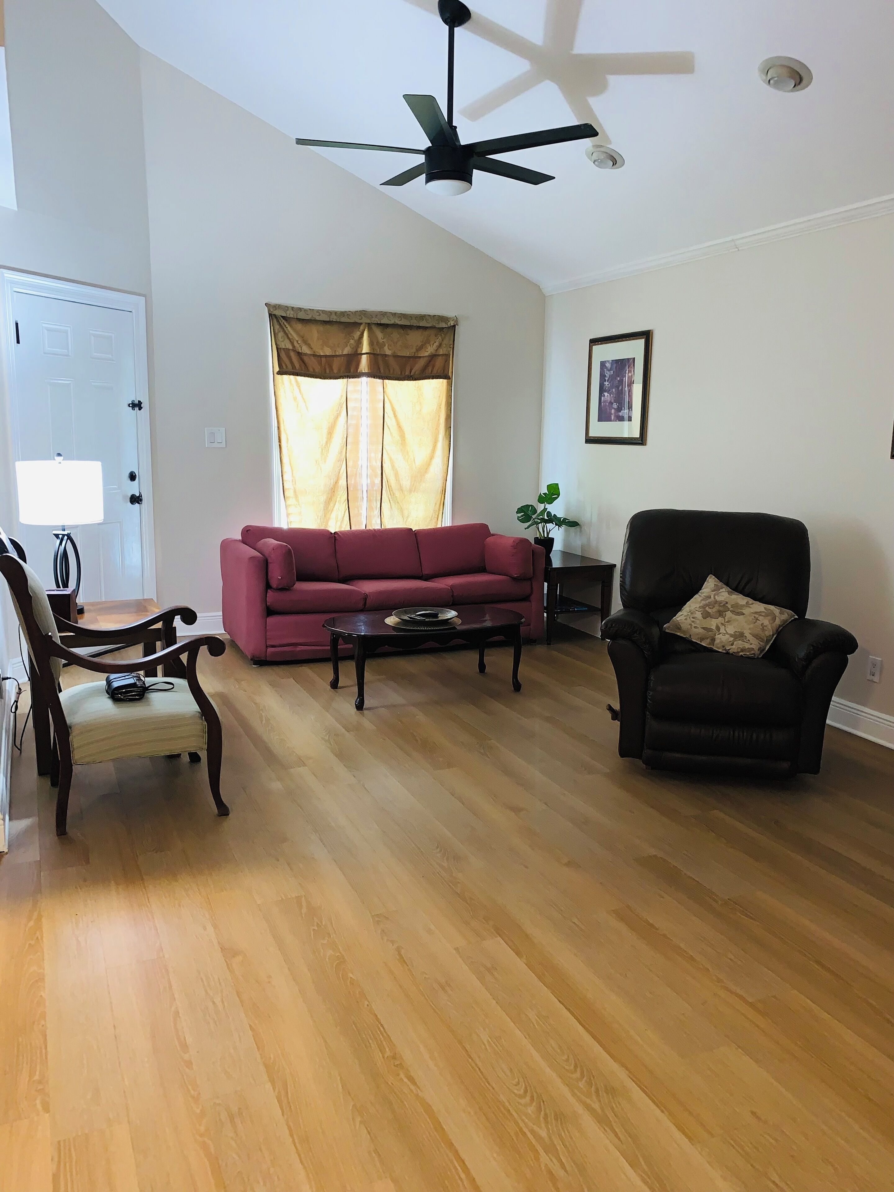 Un salon avec un plancher en bois, un canapé rouge, un fauteuil noir, un ventilateur de plafond et une fenêtre avec des rideaux.