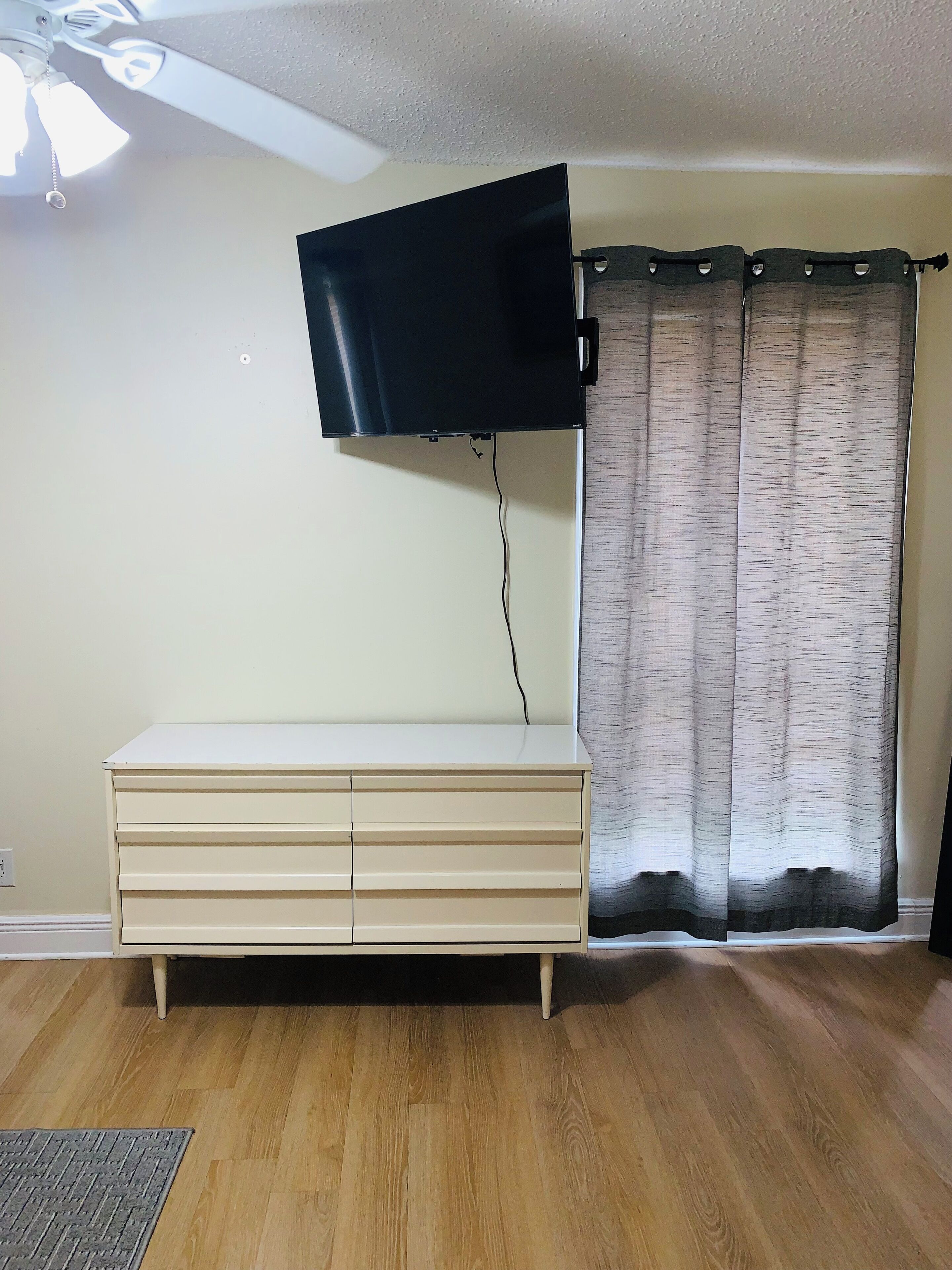 A room with a flat-screen TV mounted on the wall, a wooden cabinet, a window with curtains, and a ceiling fan.