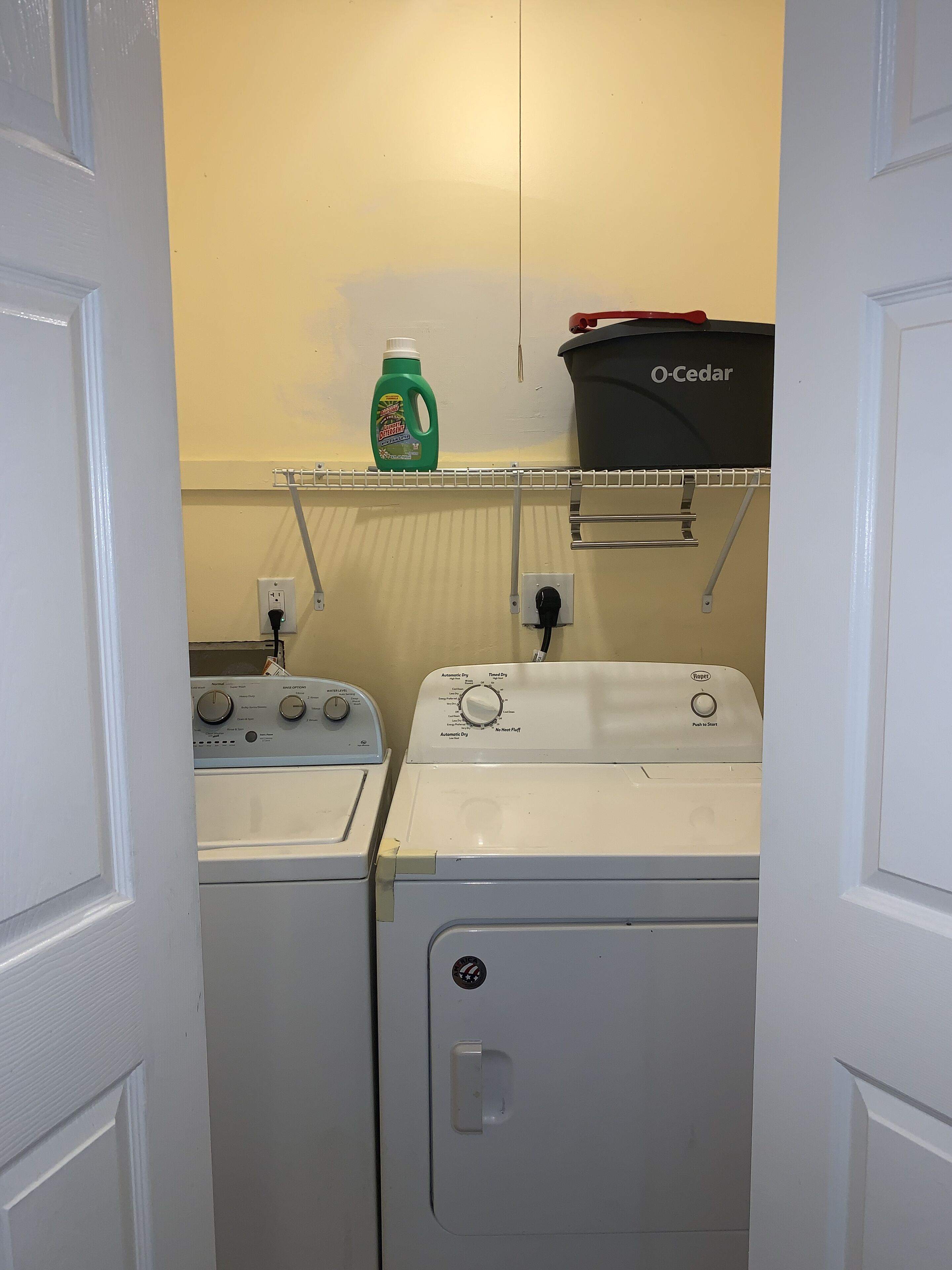 A laundry area with two washing machines, a shelf with a detergent bottle, and a trash bin.
