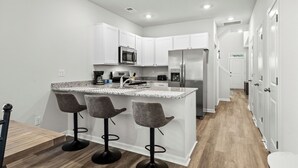 A modern kitchen with white cabinets, a stainless steel refrigerator, and a granite countertop. There are three bar stools and a microwave above the counter. - 3-bedroom townhome in amazing Calabash next to Meadowlands Golf Course & Beach (Calabash)