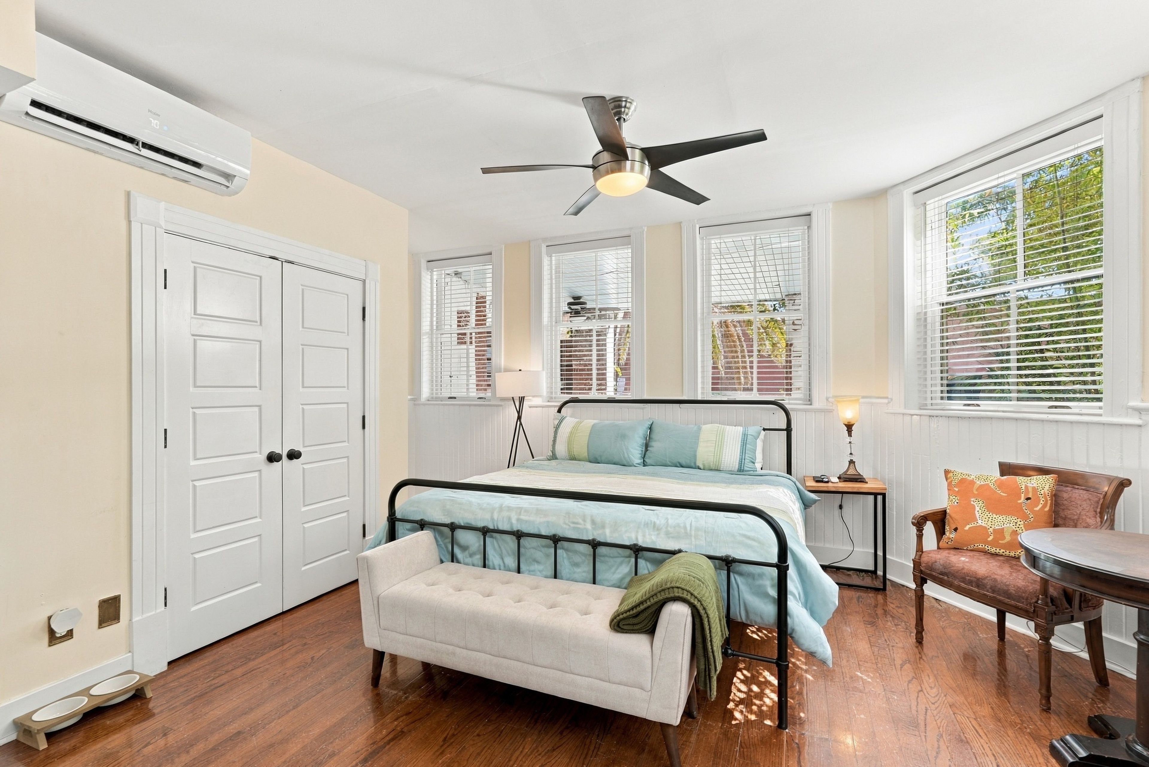 A bedroom with a bed, a bench, a chair, a table, a ceiling fan, and a window with blinds.