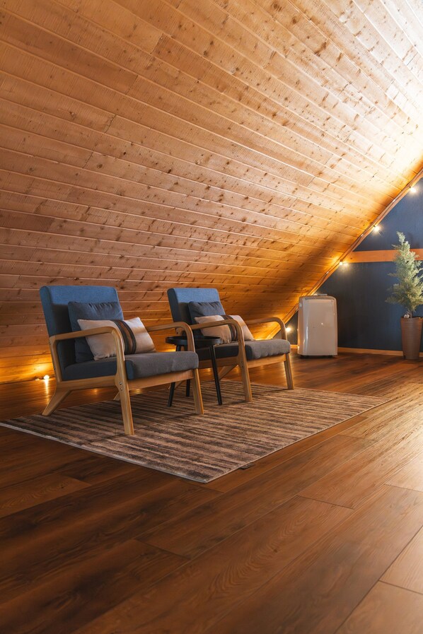 Un cuarto ático acogedor con listones de madera en el techo, dos sillones azules con cojines, un refrigerador blanco pequeño y una planta en maceta.