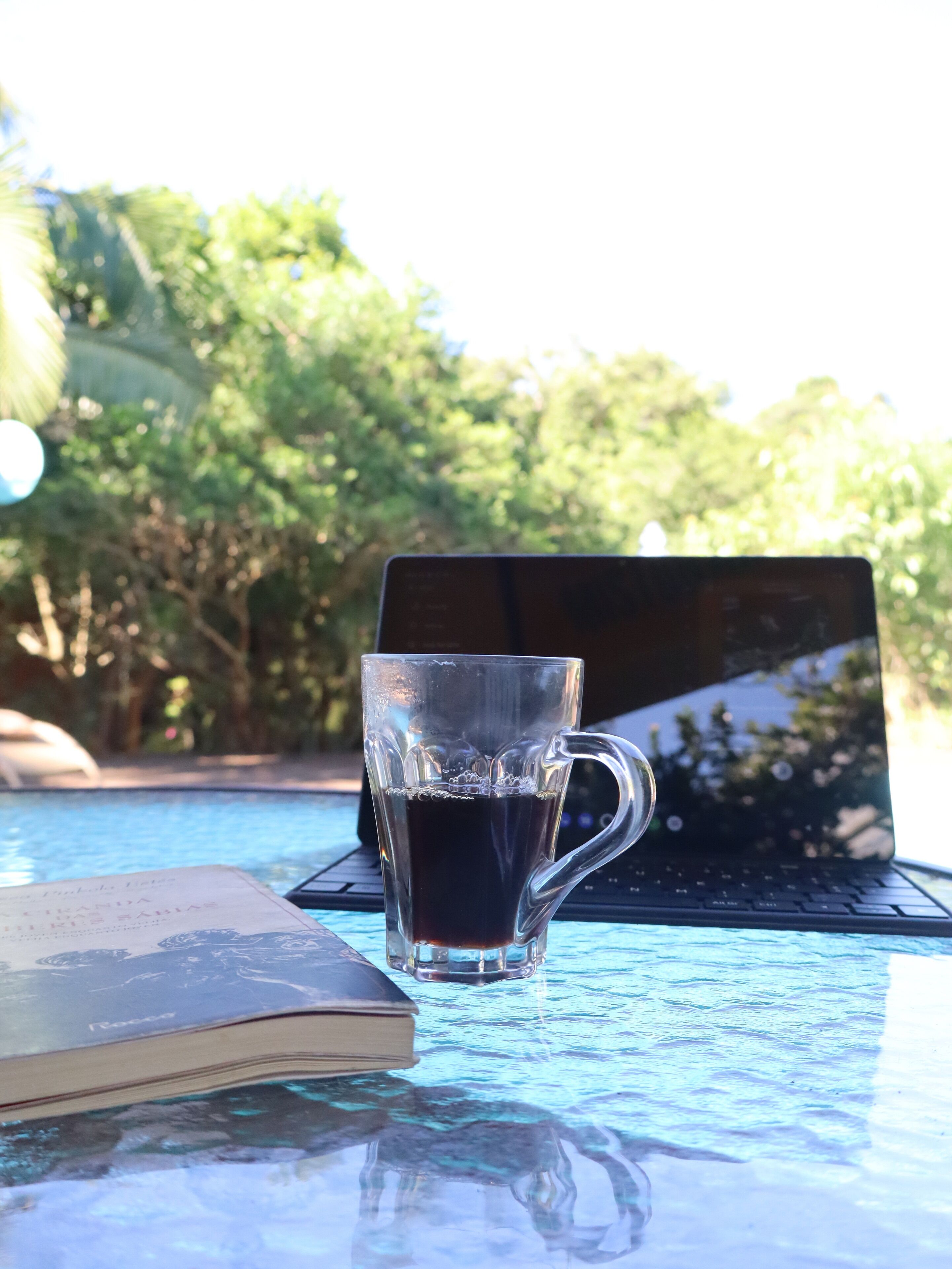 En bærbar computer på et glasbord med et klart glas og en åben bog ved siden af.