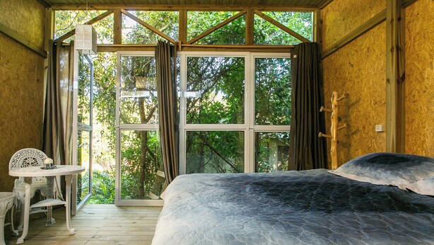 A bedroom with a large bed, a small table, and a view of trees through a glass door. - Pousada guarda beach house (Palhoca)