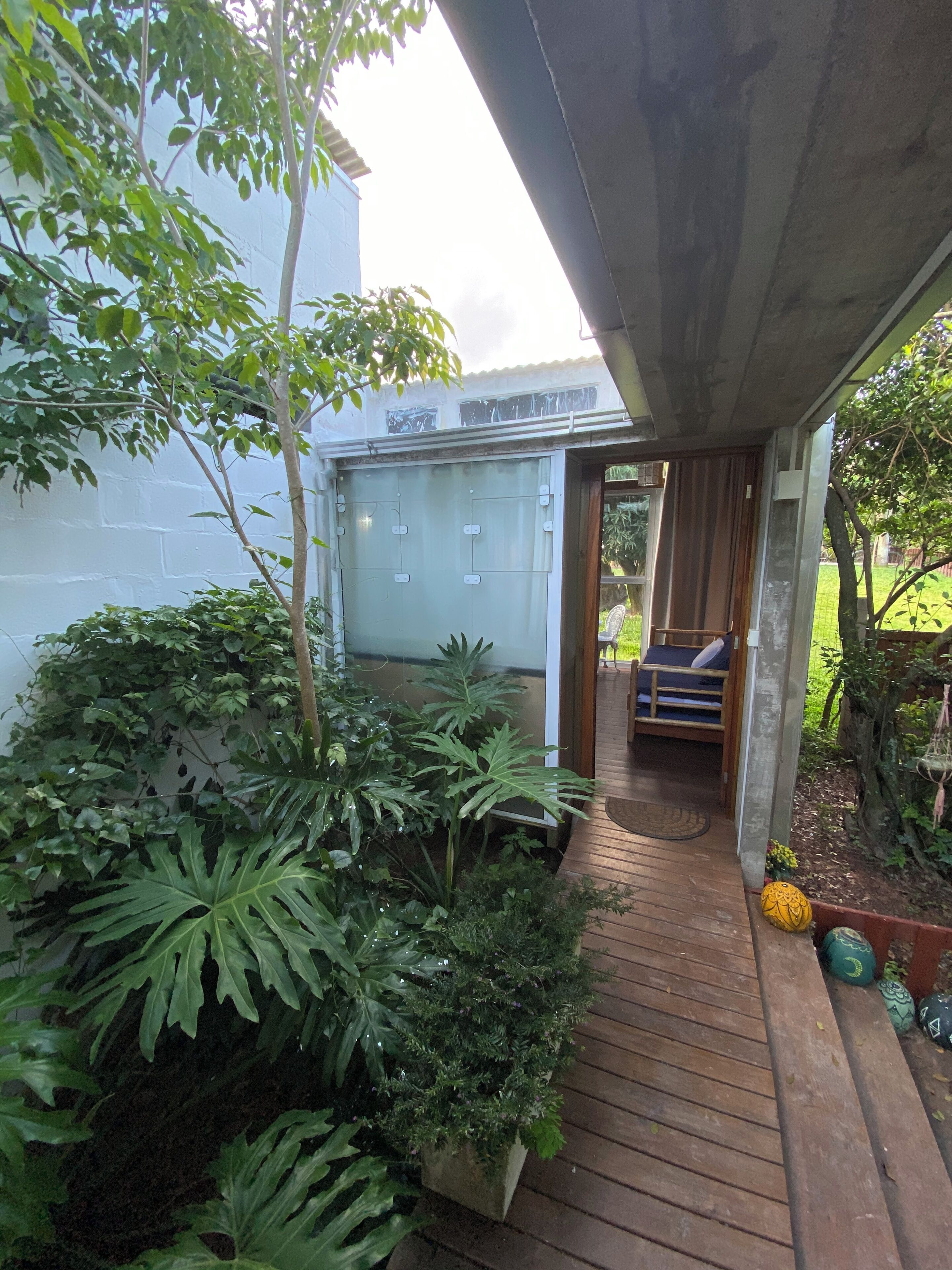 Um deck de madeira leva a uma área coberta com espaço para sentar, com vista para um jardim e um edifício ao fundo.