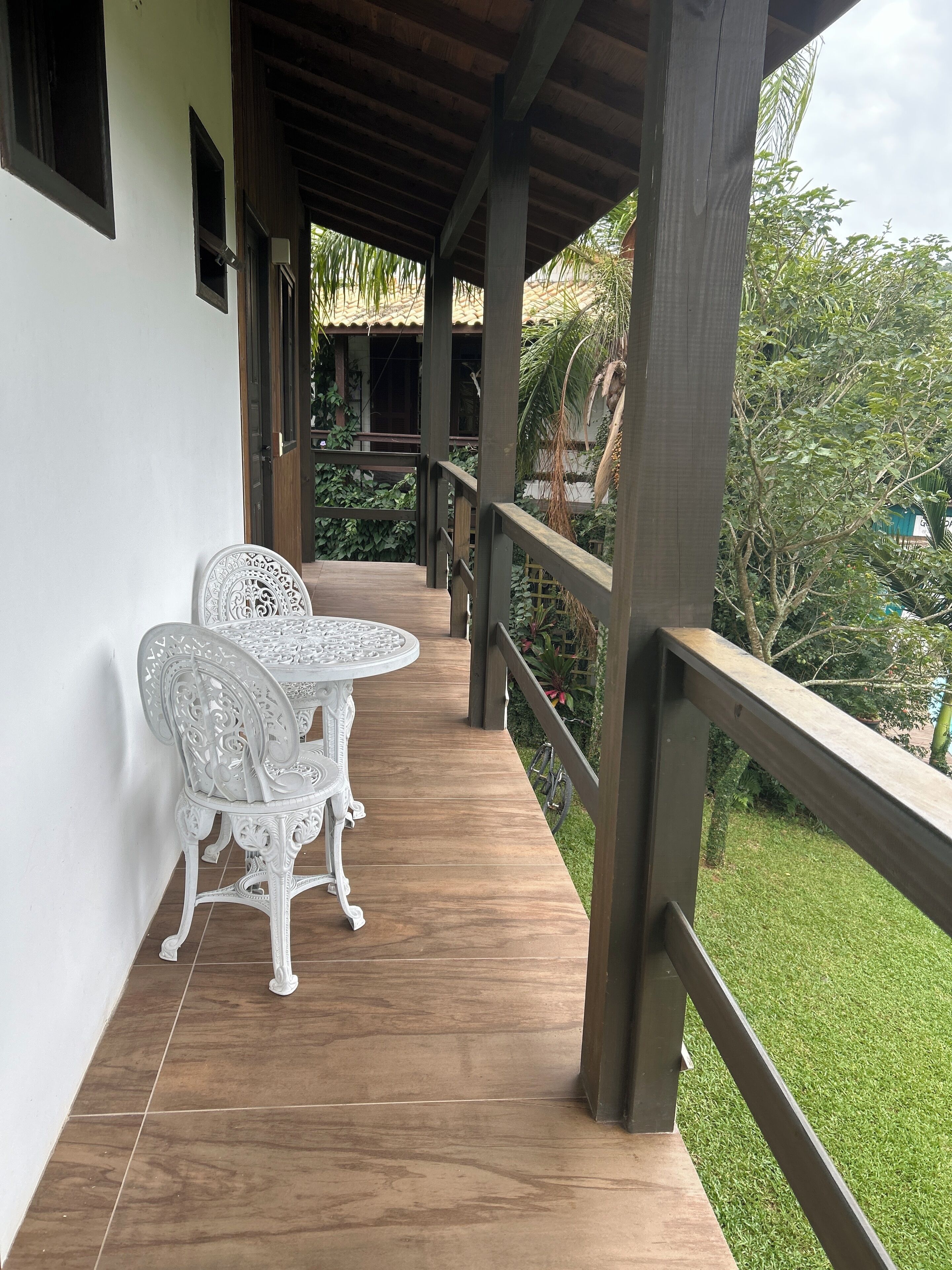 A covered balcony with a wooden floor, white ornate chairs, and a view of a garden.