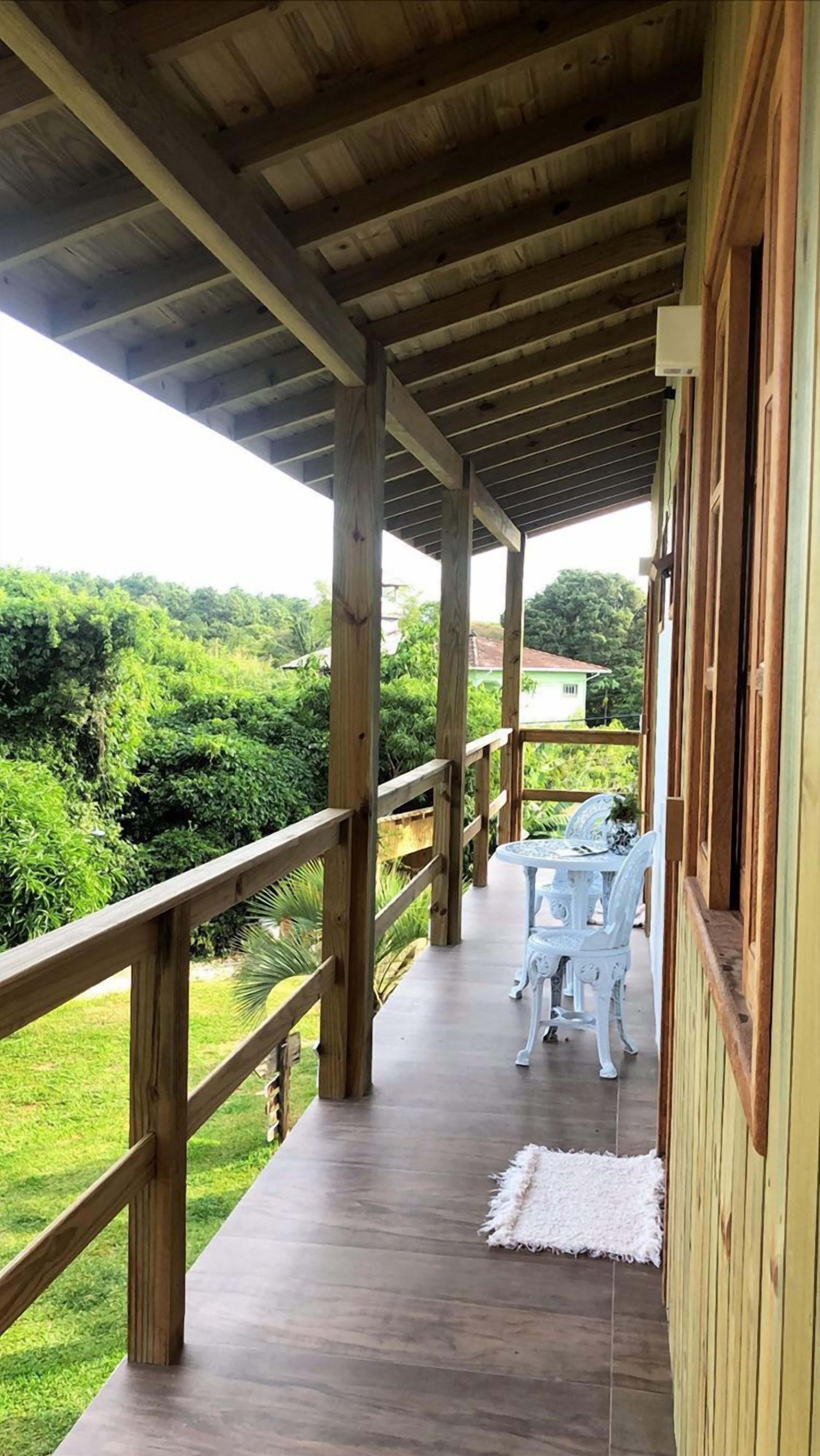 Un espacio al aire libre techado con una terraza de madera, sillas blancas y vistas a la vegetación.