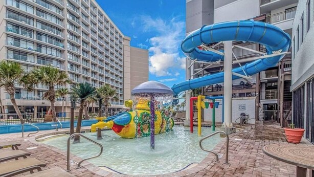 Zona de parque acuático con un tobogán gigante, una fuente de agua colorida y sillas de descanso junto a la piscina.