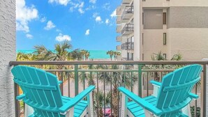 Un balcón con sillas Adirondack turquesa, con vista a una playa con palmeras y un edificio con balcones.