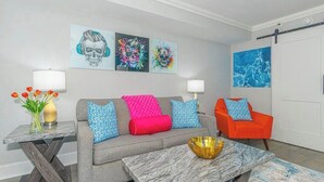 Una sala de estar con un sofá gris, cojines rojos y azules, un sillón rojo, una mesa de centro de mármol y tres obras de arte en la pared.